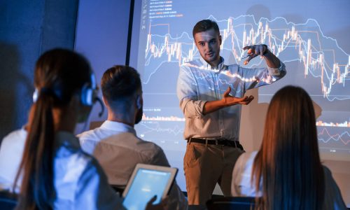 Company growth plan. Male leader talking to employees, showing information on the projector in office of stock exchange company.